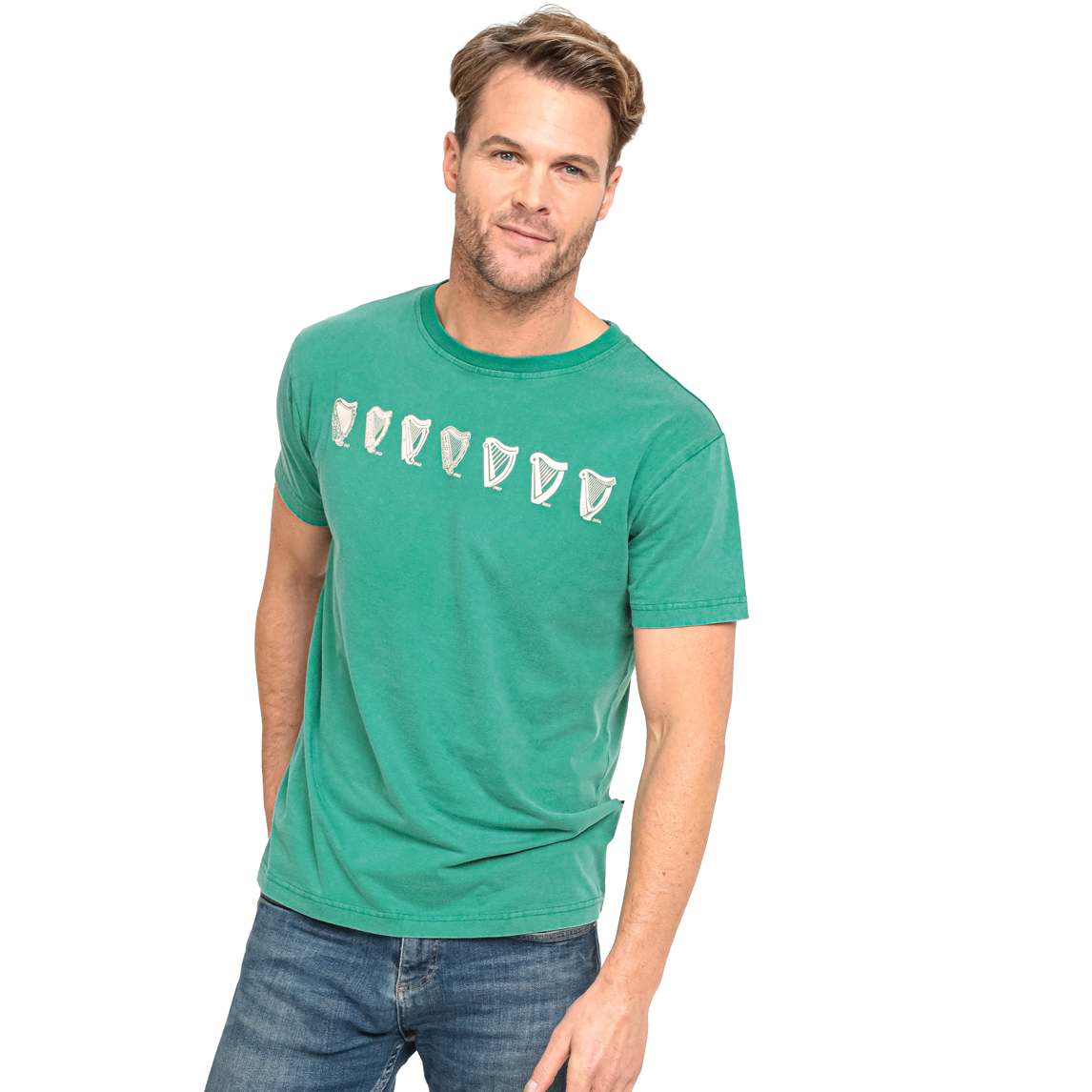A man with short hair is wearing a blue jeans and a Guinness Green Evolution Harp Premium Tee, featuring multiple white harp symbols across the chest, standing against a plain white background.