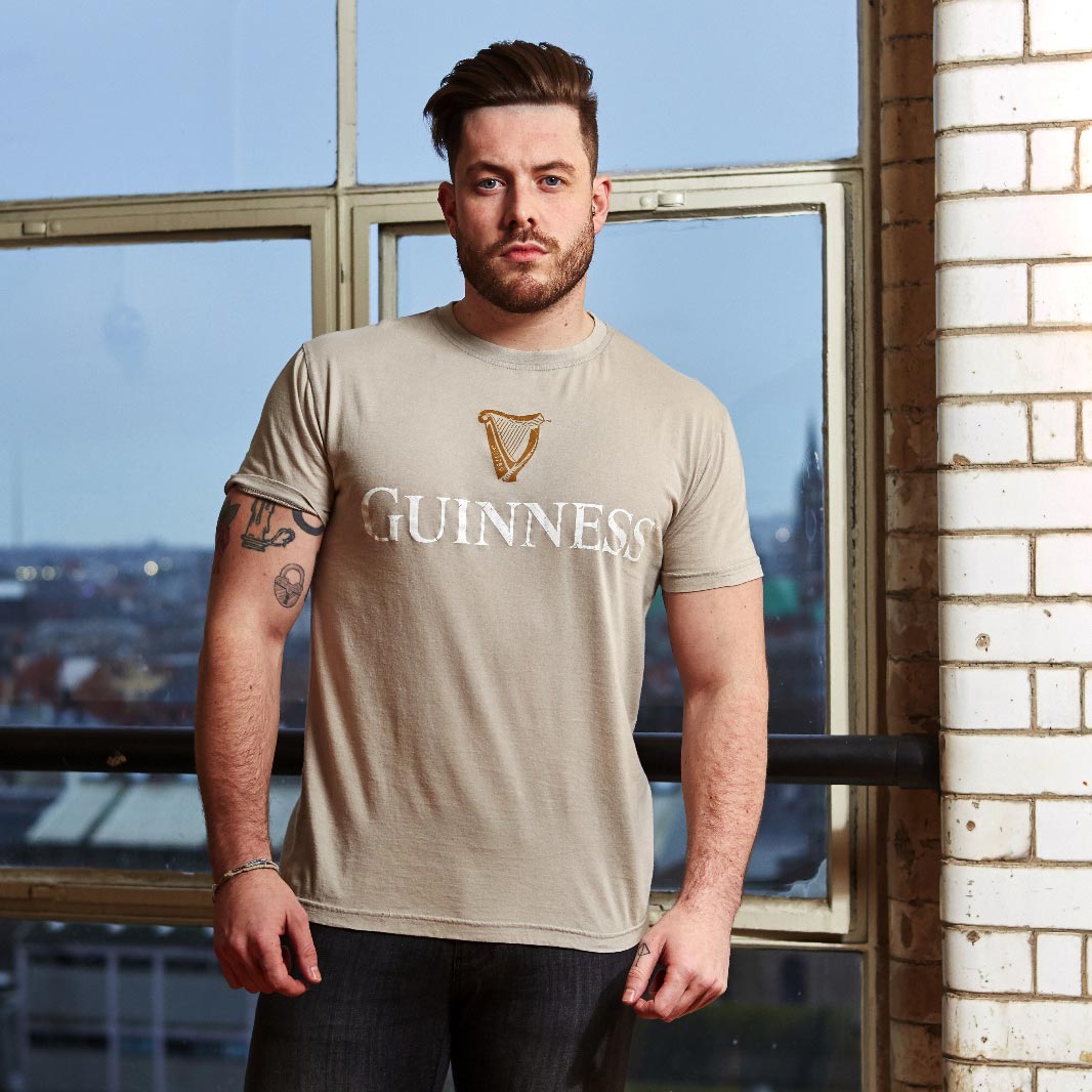 A man with short hair and a beard stands indoors in front of a window, wearing a Beige Guinness Harp Premium Tee made from 100% cotton and jeans.