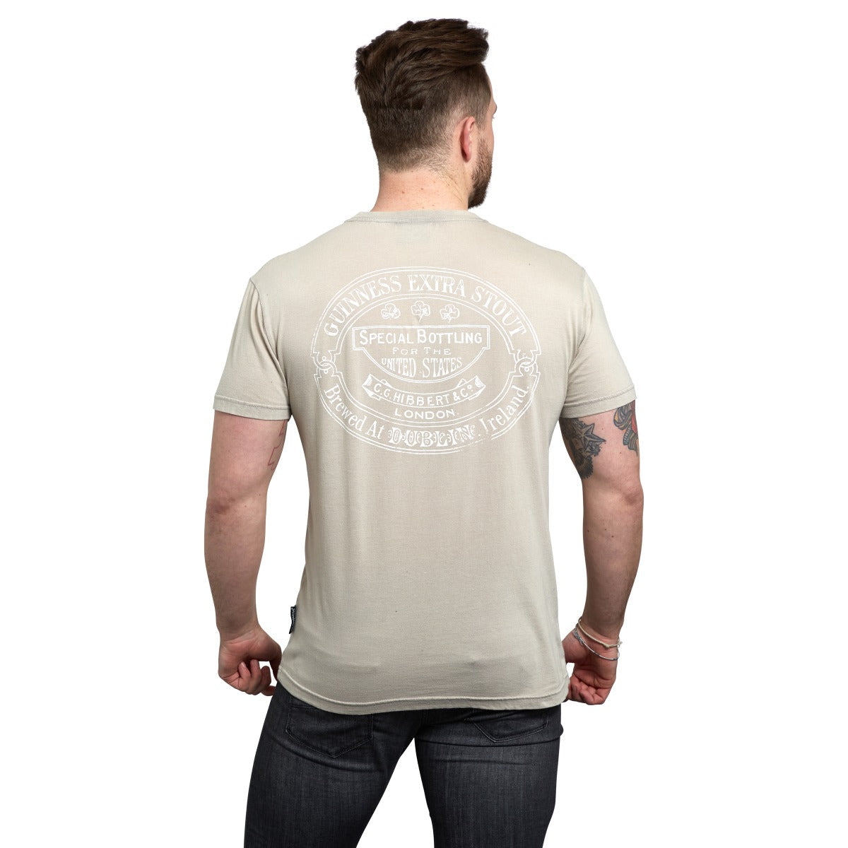 A person with short hair and a beard is facing away, wearing a beige Guinness Harp Premium Tee by Guinness with a circular design and text on the back, along with dark pants.