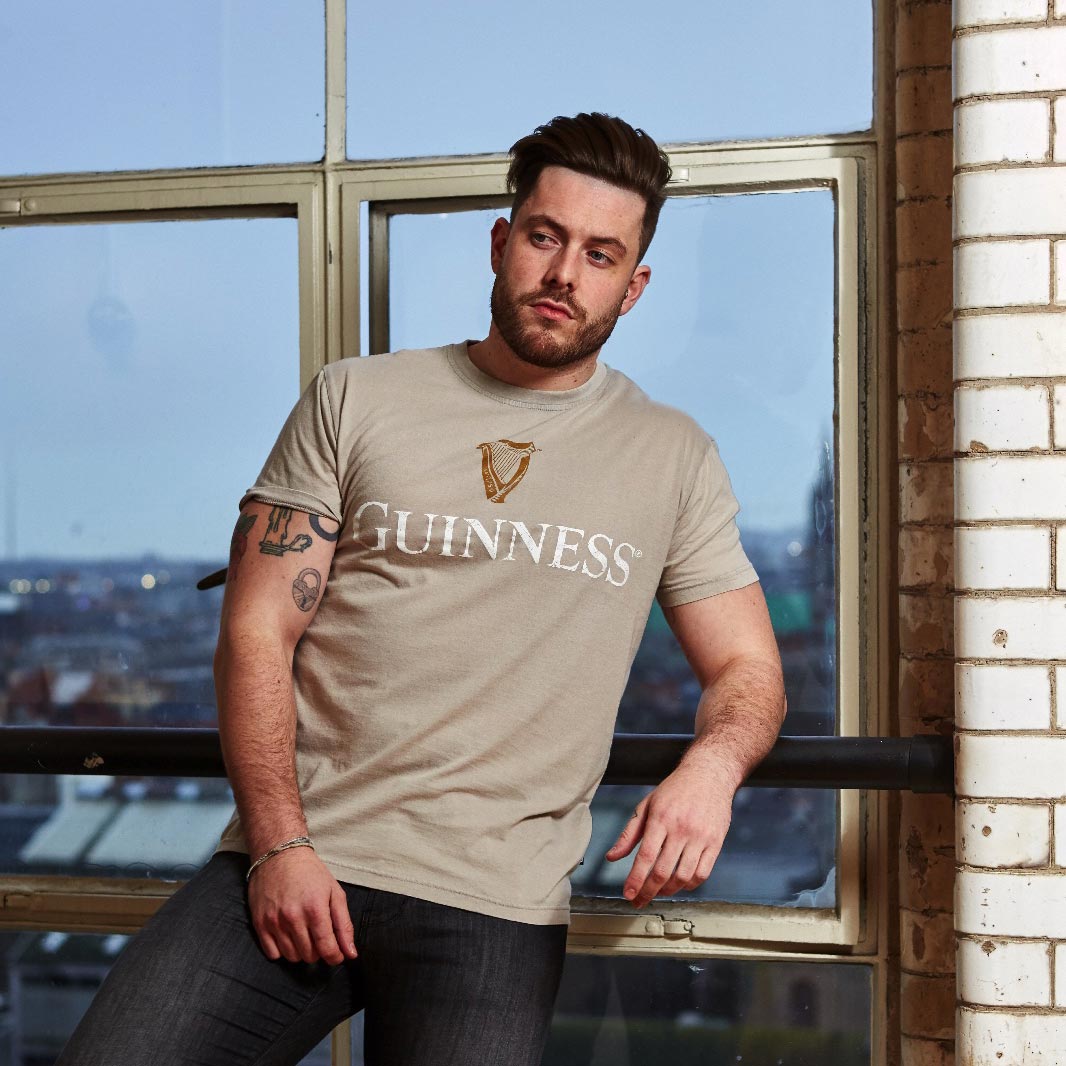 A man wearing a beige Guinness Harp Premium Tee stands by a window with a cityscape in the background.