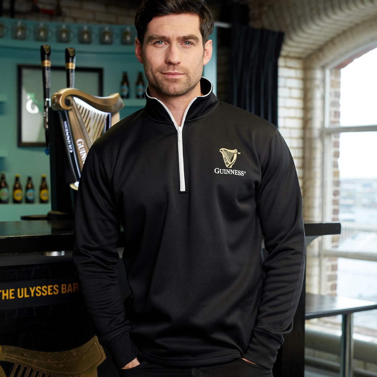 A fan wearing a Guinness Harp Half Zip Sweater is standing in front of a bar.