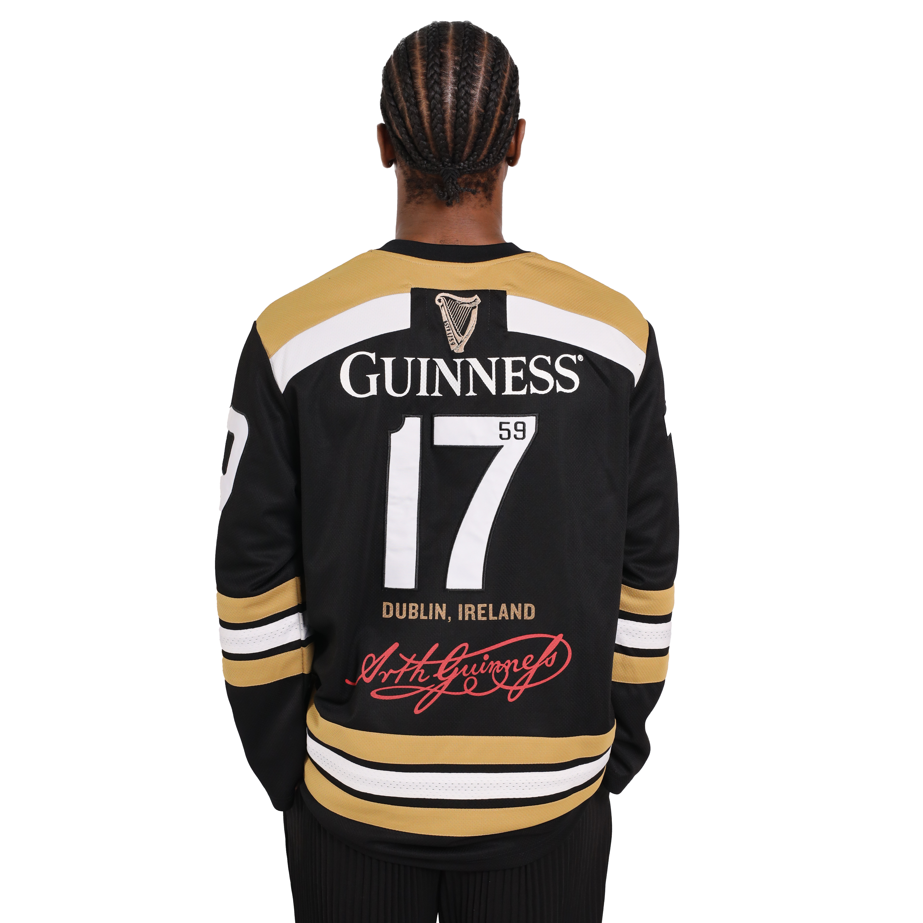 A person with braided hair wearing a black and gold Guinness Webstore US Guinness Tortoise Hockey Jersey, featuring the number 17 and the text "Guinness 1759 Dublin, Ireland" on the back, stands facing away. This sporty design is crafted from recycled polyester for an eco-friendly twist.