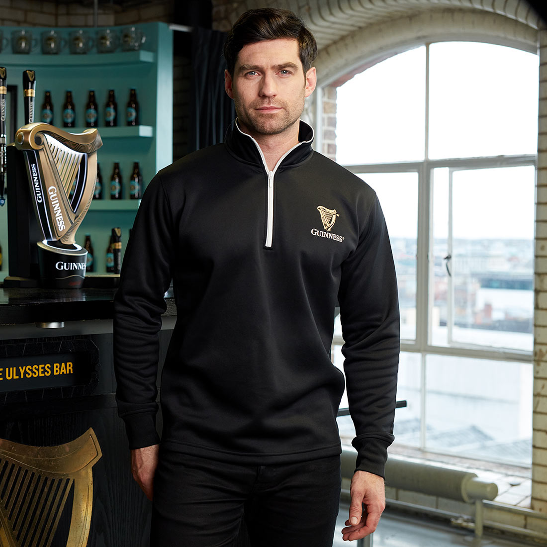A Guinness fan standing in front of a bar wearing a Guinness Harp Half Zip Sweater.
