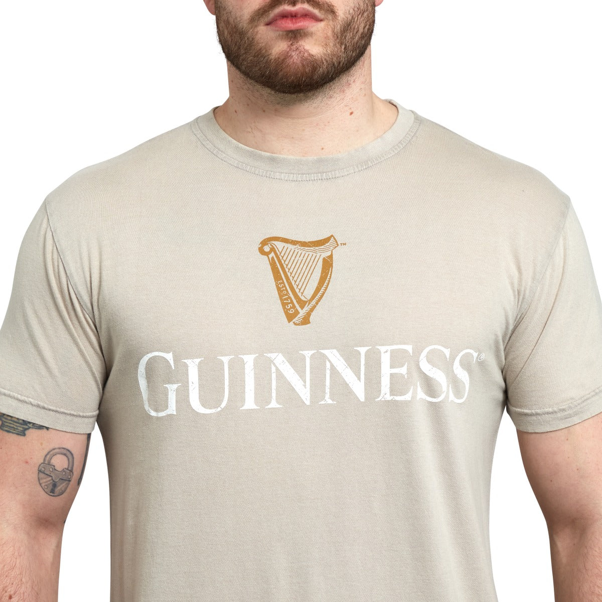 A person wearing a beige Guinness Harp Premium Tee with the iconic logo and emblem printed on the front, made from 100% cotton.