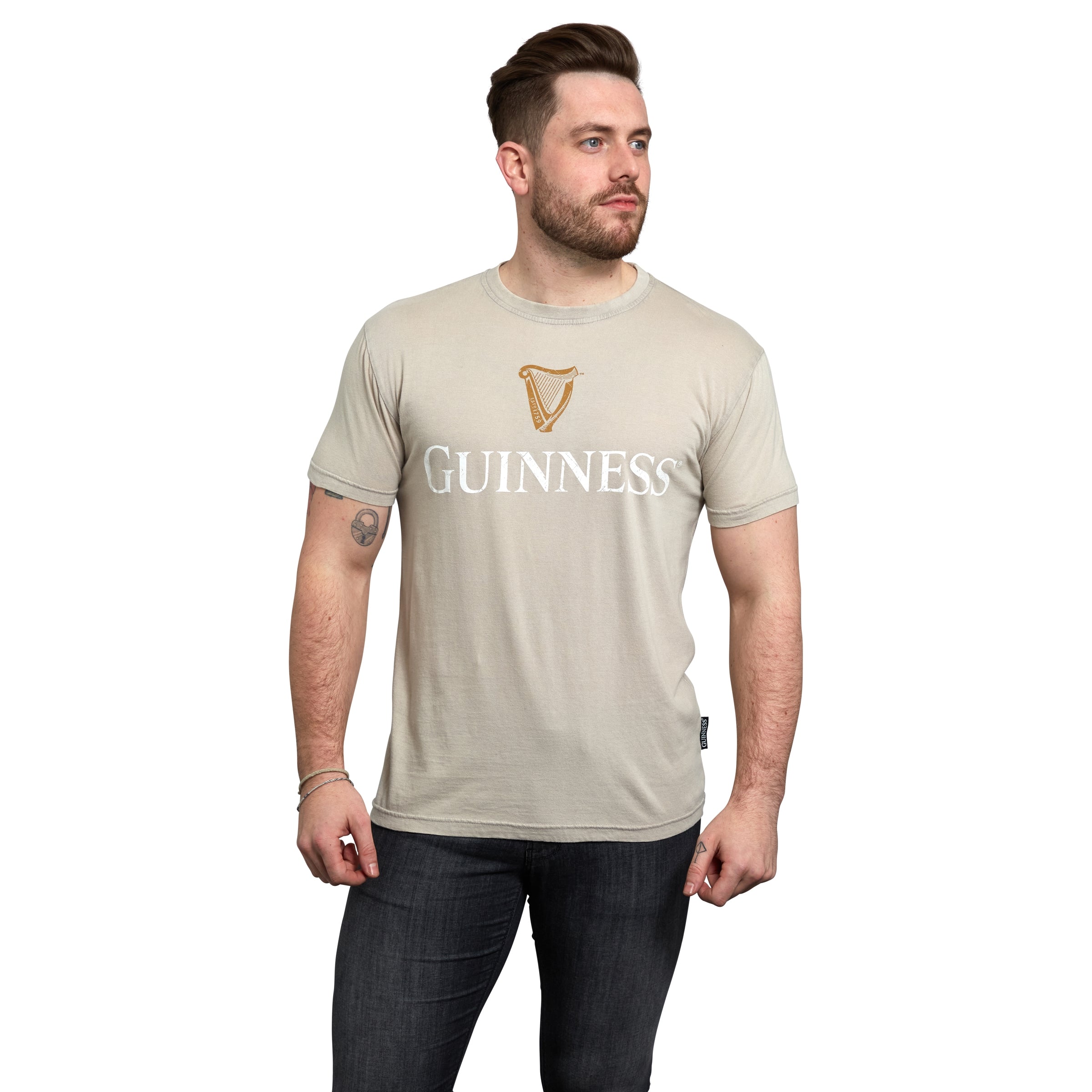 A man wearing a beige Guinness Harp Premium Tee with a trademark label stands with hands by his sides, looking off to the left.