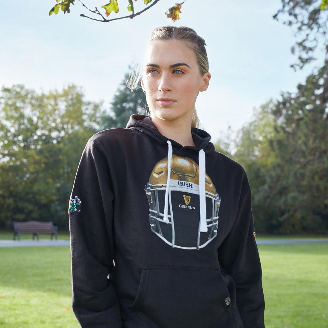 A woman celebrating in comfort, wearing a Guinness Notre Dame Helmet Hoodie Black with a brand logo gold helmet on it.