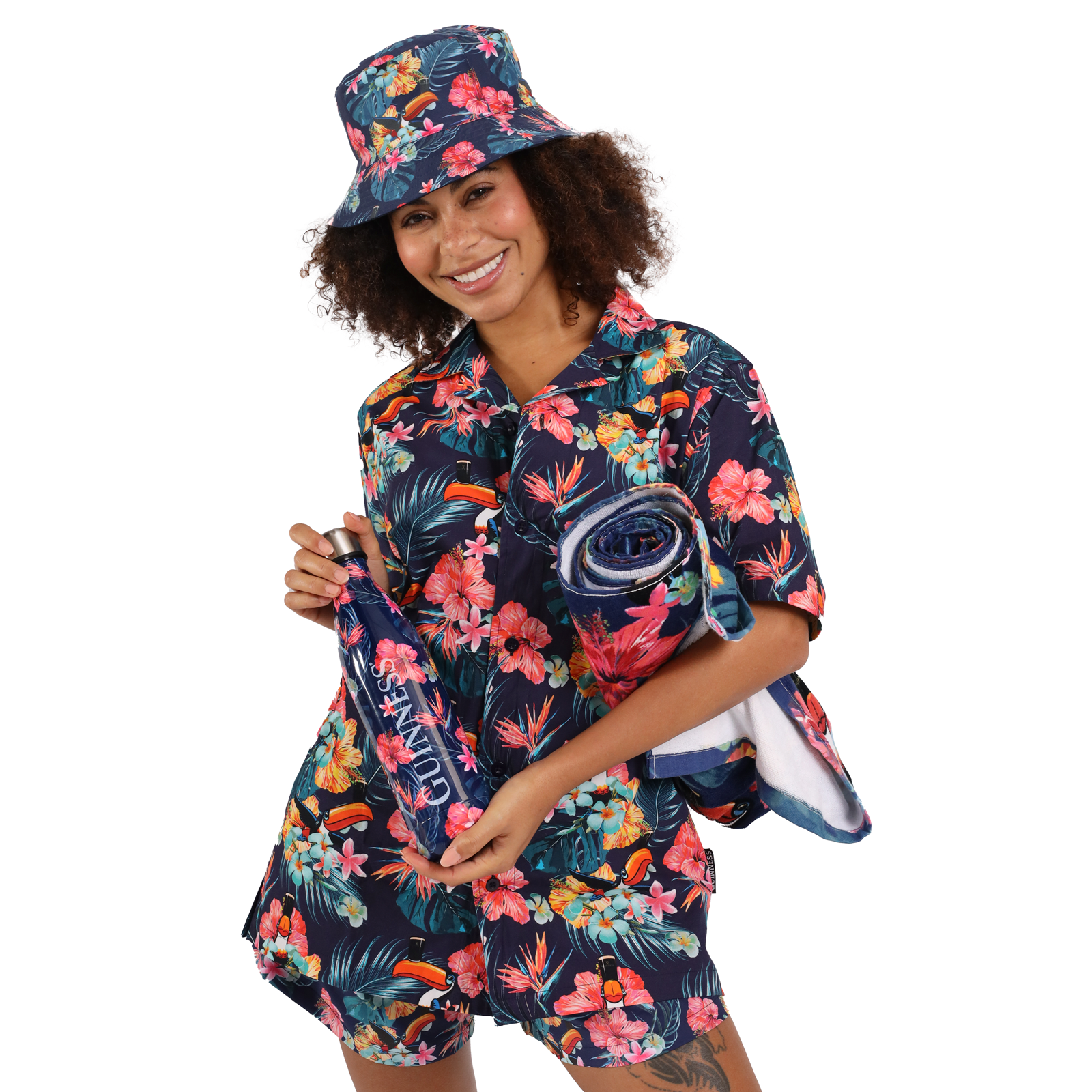 Person smiling and wearing a matching floral-patterned shirt, shorts, and bucket hat, holding a rolled-up towel and a small bag with a Guinness Toucan Hawaiian Water Bottle by Guinness peeking out.