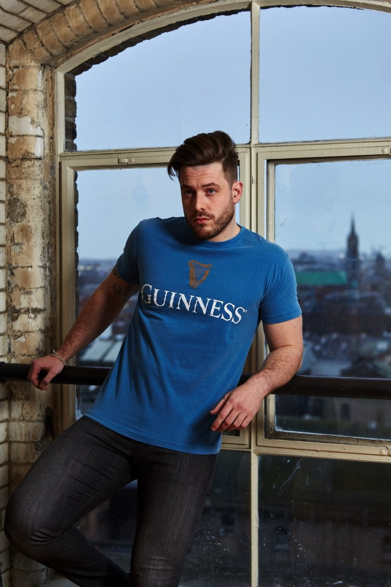 A man wearing a Blue Guinness Harp Premium Tee t-shirt leans against a windowsill in a room with brick walls, with a view of buildings outside the window.