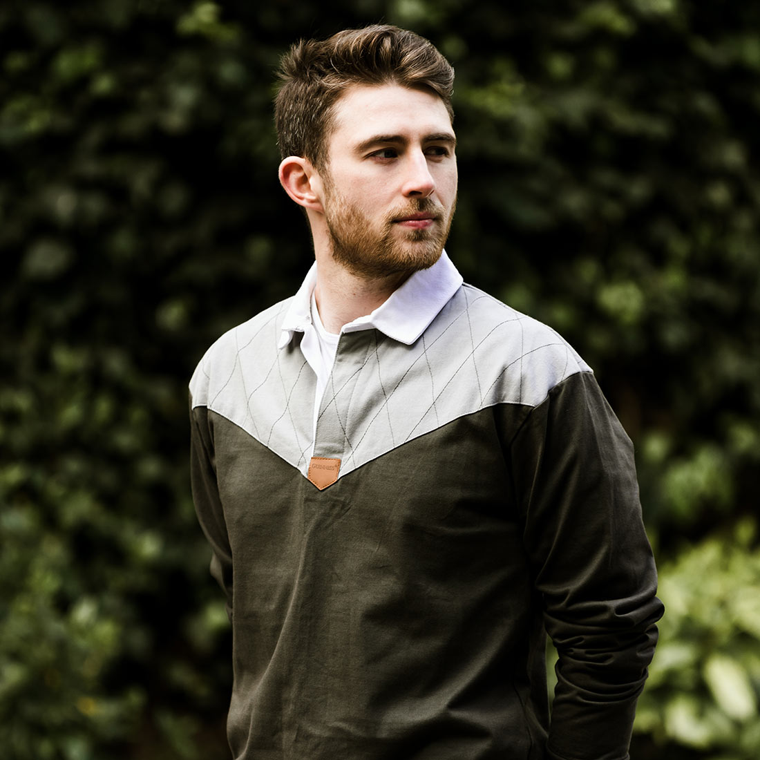 A man in a Guinness Heritage Charcoal Grey & Black Long-Sleeve Rugby jersey is standing in front of bushes.