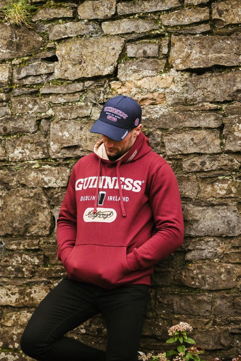 A man wearing a burgundy hoodie and a Guinness Signature Blue Baseball Cap Adjustable is leaning against a stone wall.