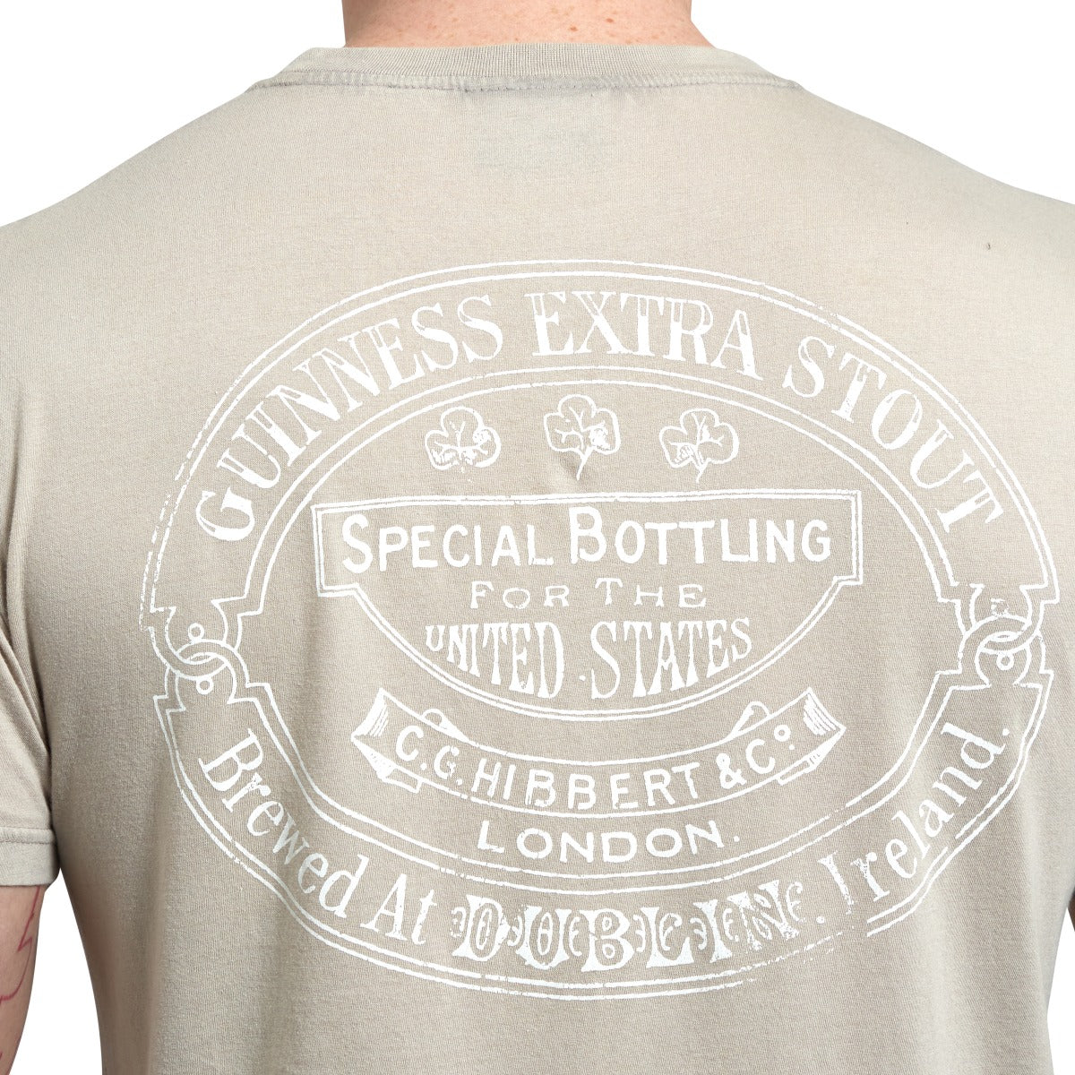 Back view of a person wearing a 100% cotton beige Guinness Harp Premium Tee with a white printed design that reads "Guinness Extra Stout," "Special Bottling for the United States," and other details, including "Dublin" and "London.