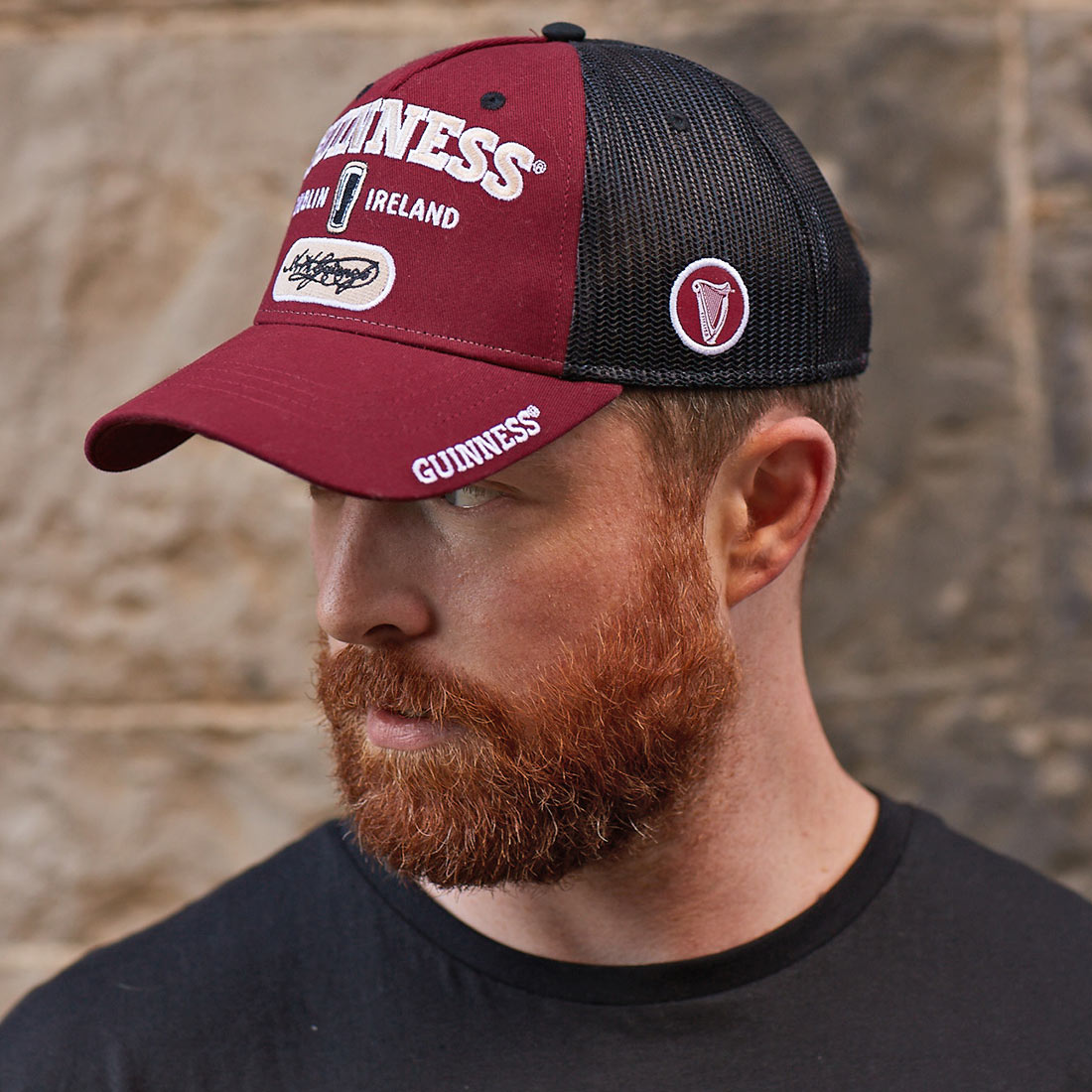 A bearded man wearing a Guinness Signature Burgundy Trucker Mesh Baseball Cap Adjustable.