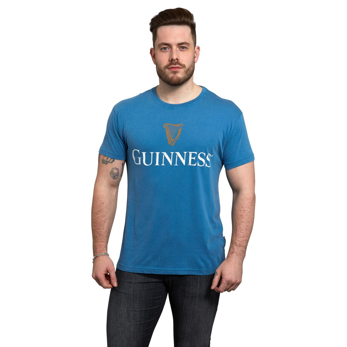 A person wearing a Blue Guinness Harp Premium Tee with the Guinness® Trademark Label and dark jeans, standing against a plain white background.