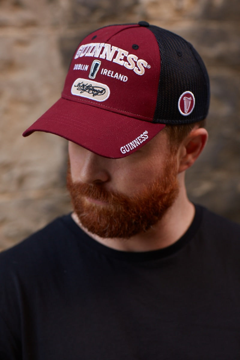 A man with a red beard wearing a Guinness Signature Burgundy Trucker Mesh Baseball Cap Adjustable.