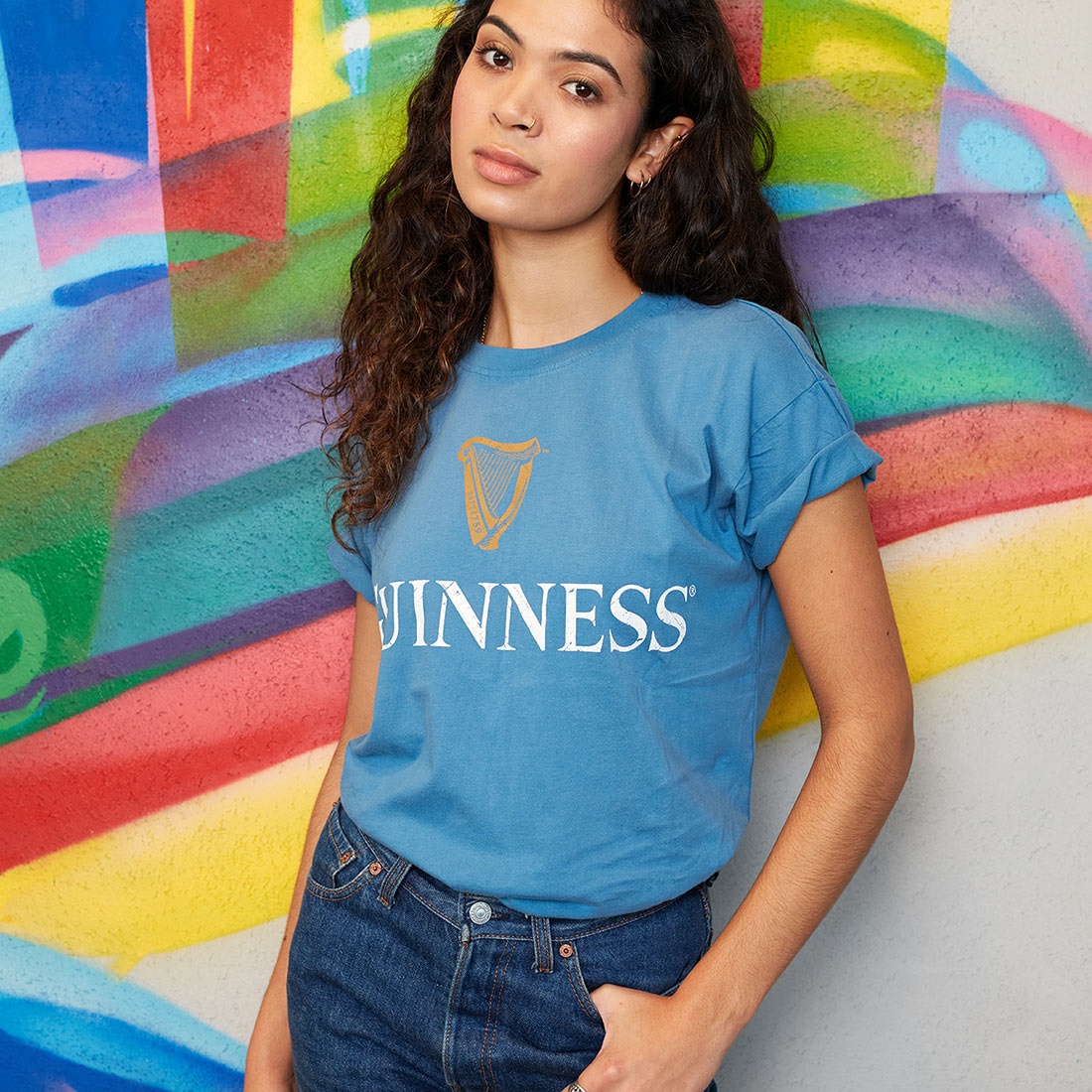 A woman with long curly hair wearing a Blue Guinness Harp Premium Tee and jeans stands against a colorful graffiti wall, looking at the camera with one hand in her pocket.