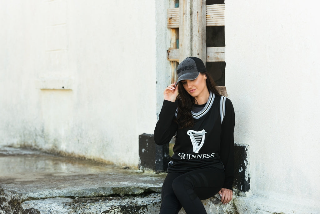 A woman sitting on a stone wall wearing a Guinness Trucker Hat by Guinness.