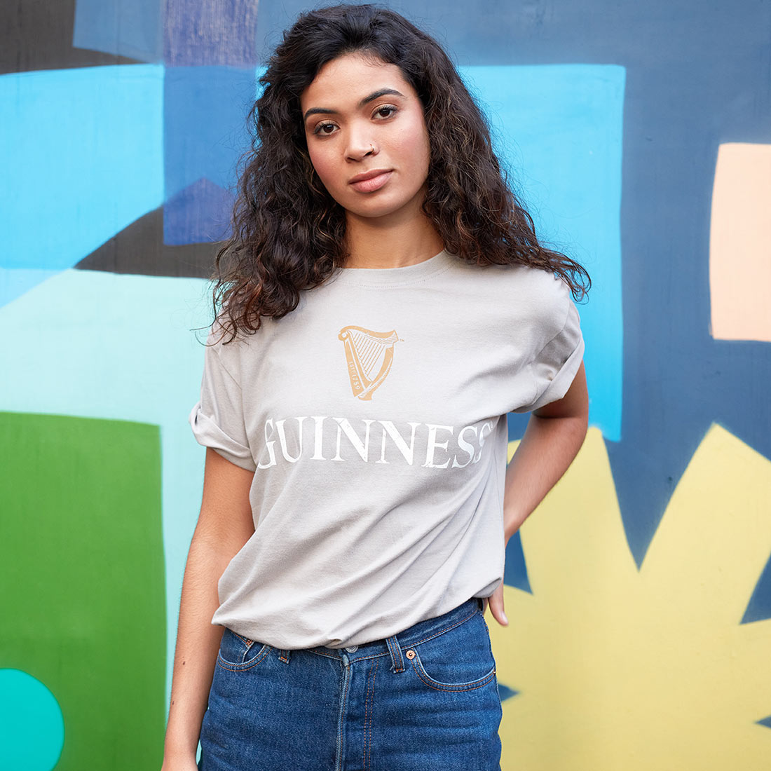 A young woman with wavy hair wearing a beige "Guinness Harp Premium Tee" and jeans stands in front of a colorful geometric mural.