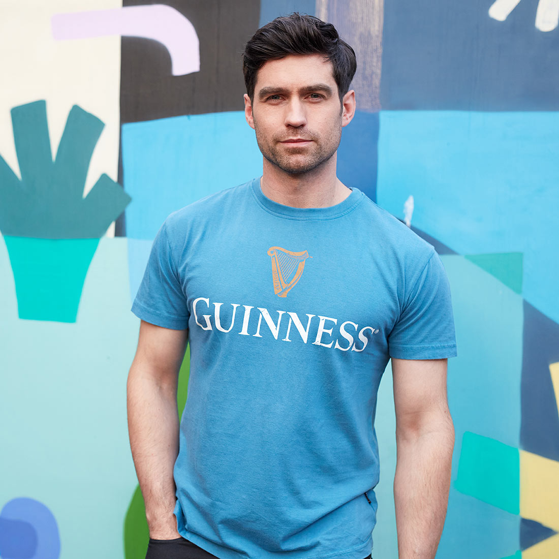 A man wearing a vintage Blue Guinness Harp Premium Tee stands in front of a colorful abstract mural, looking at the camera.