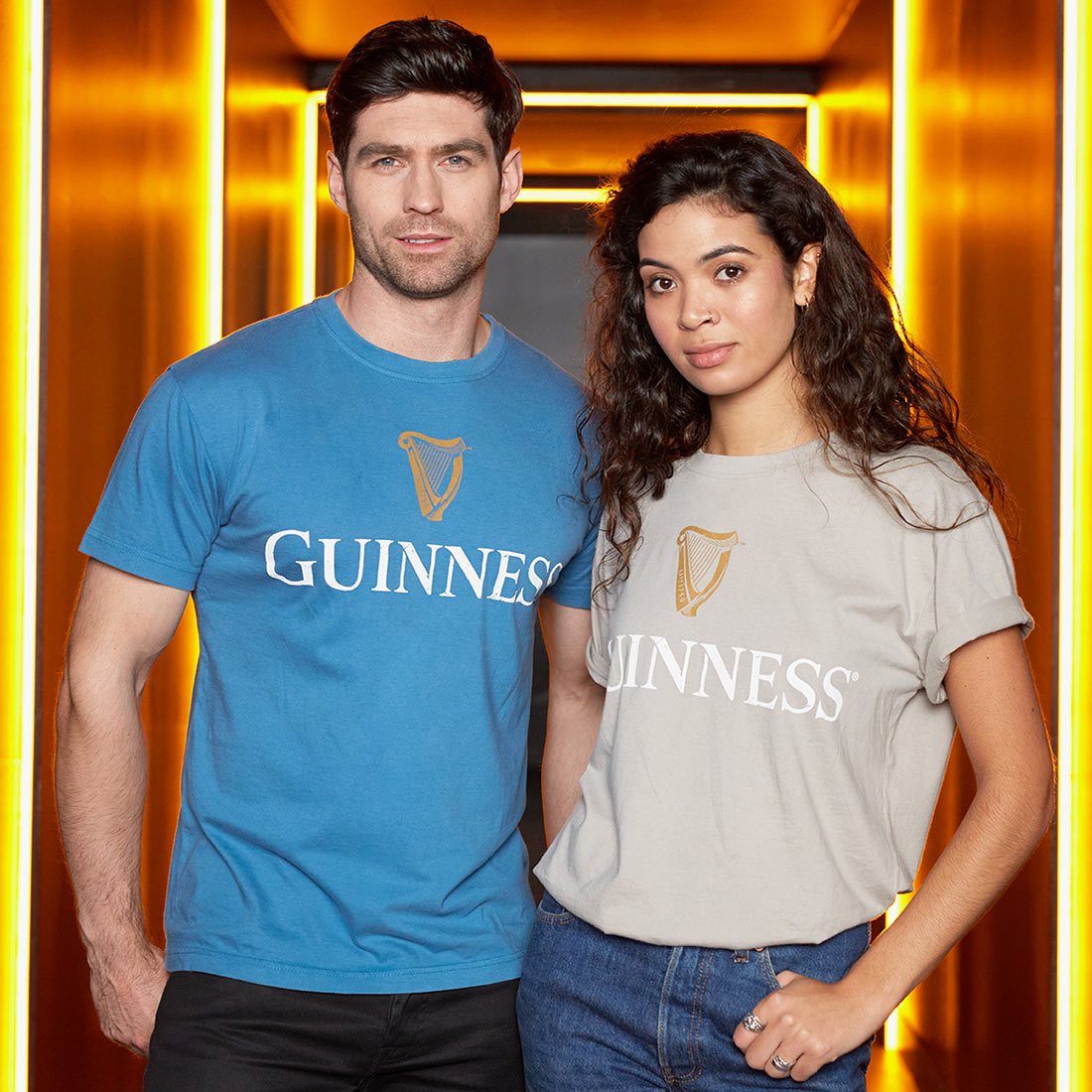 Two people stand side by side, one in a blue 100% cotton Guinness T-shirt and the other in a Beige Guinness Harp Premium Tee with a trademark label, in a brightly-lit area with illuminated vertical light strips.