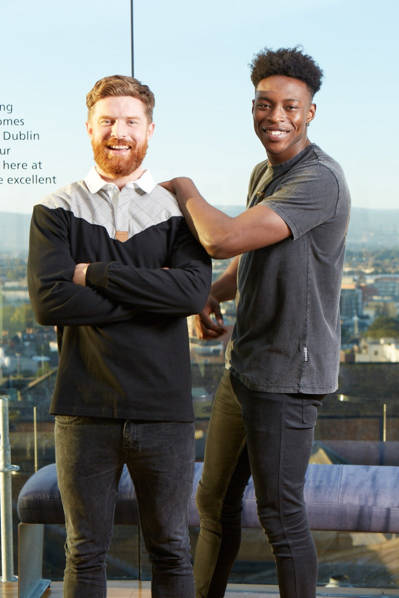 Two men wearing Guinness Heritage Charcoal Grey & Black Long-Sleeve Rugby jerseys standing next to each other in front of a building.