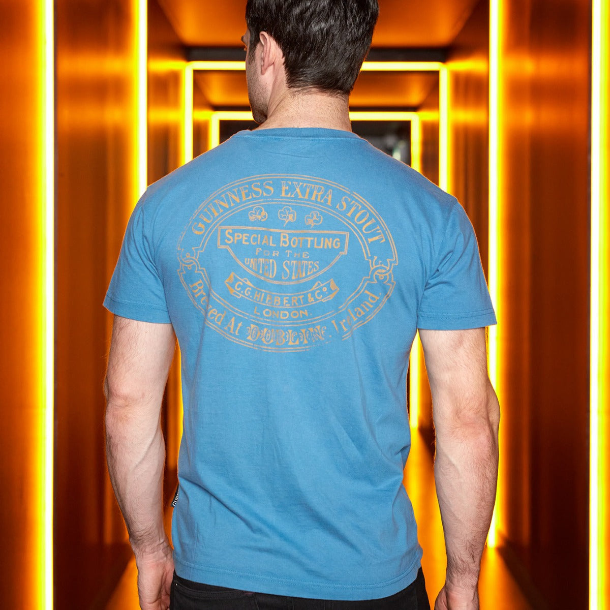 A man stands with his back to the camera in a dimly lit corridor, wearing a Blue Guinness Harp Premium Tee by Guinness.