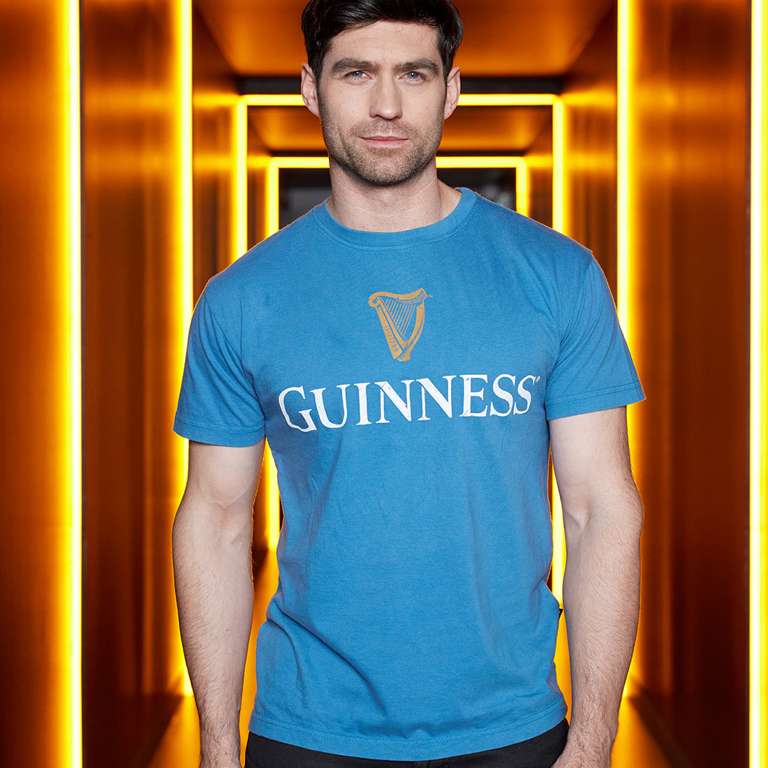 A man in a Blue Guinness Harp Premium Tee stands in a neon-lit corridor.