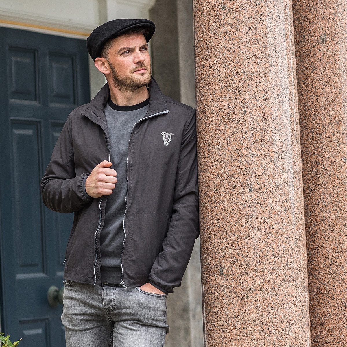 A man wearing a Guinness® Classic Felt Ivy Cap and jacket leaning against a column.