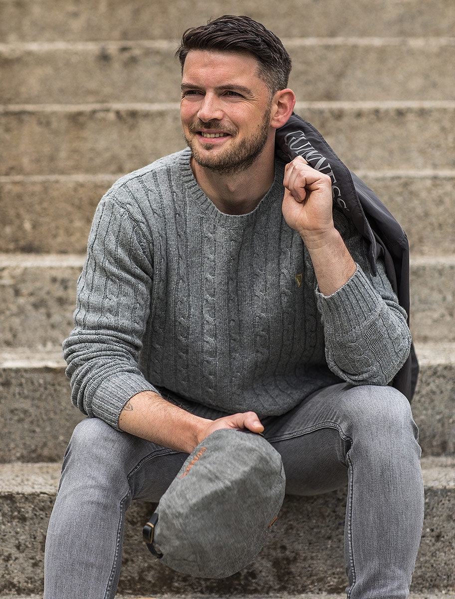 A man sitting on some steps wearing a sweater, a hat, and a Guinness Classic Tweed Buckle Ivy Flat Cap, a wardrobe essential.