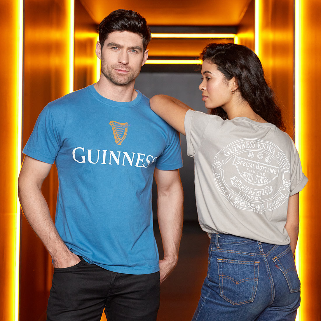 A man and woman are posing in a hallway illuminated with orange lights, both wearing vintage Guinness-themed shirts. The man is in a blue Blue Guinness Harp Premium Tee, while the woman sports a gray shirt with the large white Guinness® Trademark Label on the back.