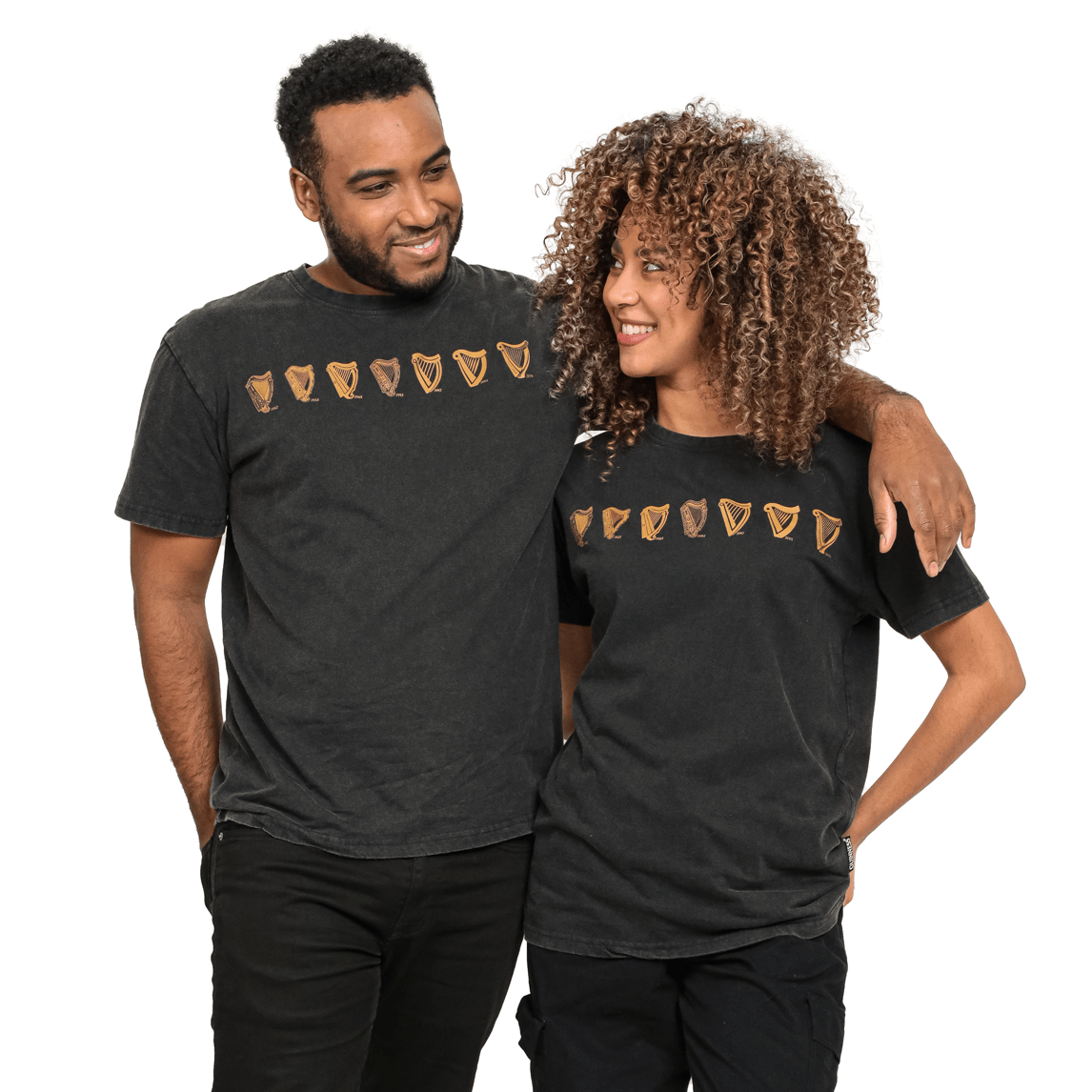 Two people wearing Guinness Black Evolution Harp Premium Tees with a line of small golden heart and harp symbols stand together, smiling and with arms around each other.