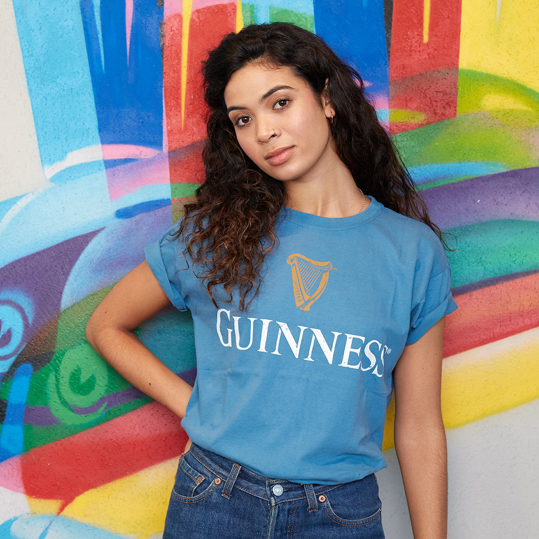 A person with long curly hair wears a Blue Guinness Harp Premium Tee featuring the Guinness® Trademark Label and stands in front of a colorful graffiti-style wall.