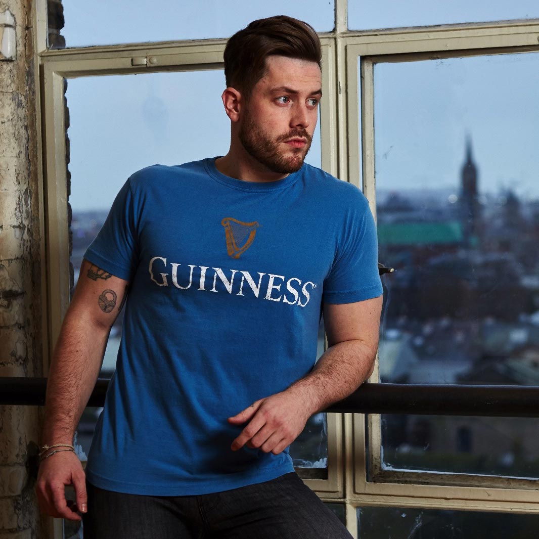 A man wearing a vintage Blue Guinness Harp Premium Tee by Guinness stands by a window with a cityscape in the background.