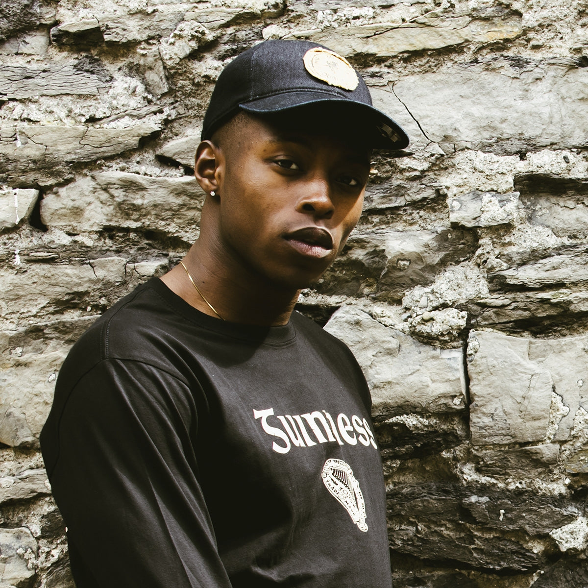 A young man wearing a black t-shirt and a Guinness Extra Stout Label baseball cap leaning against a stone wall.