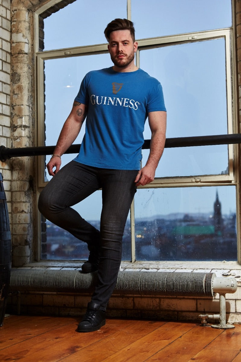 A person stands in front of a large window wearing a Blue Guinness Harp Premium Tee by Guinness and dark jeans, with a cityscape visible in the background.