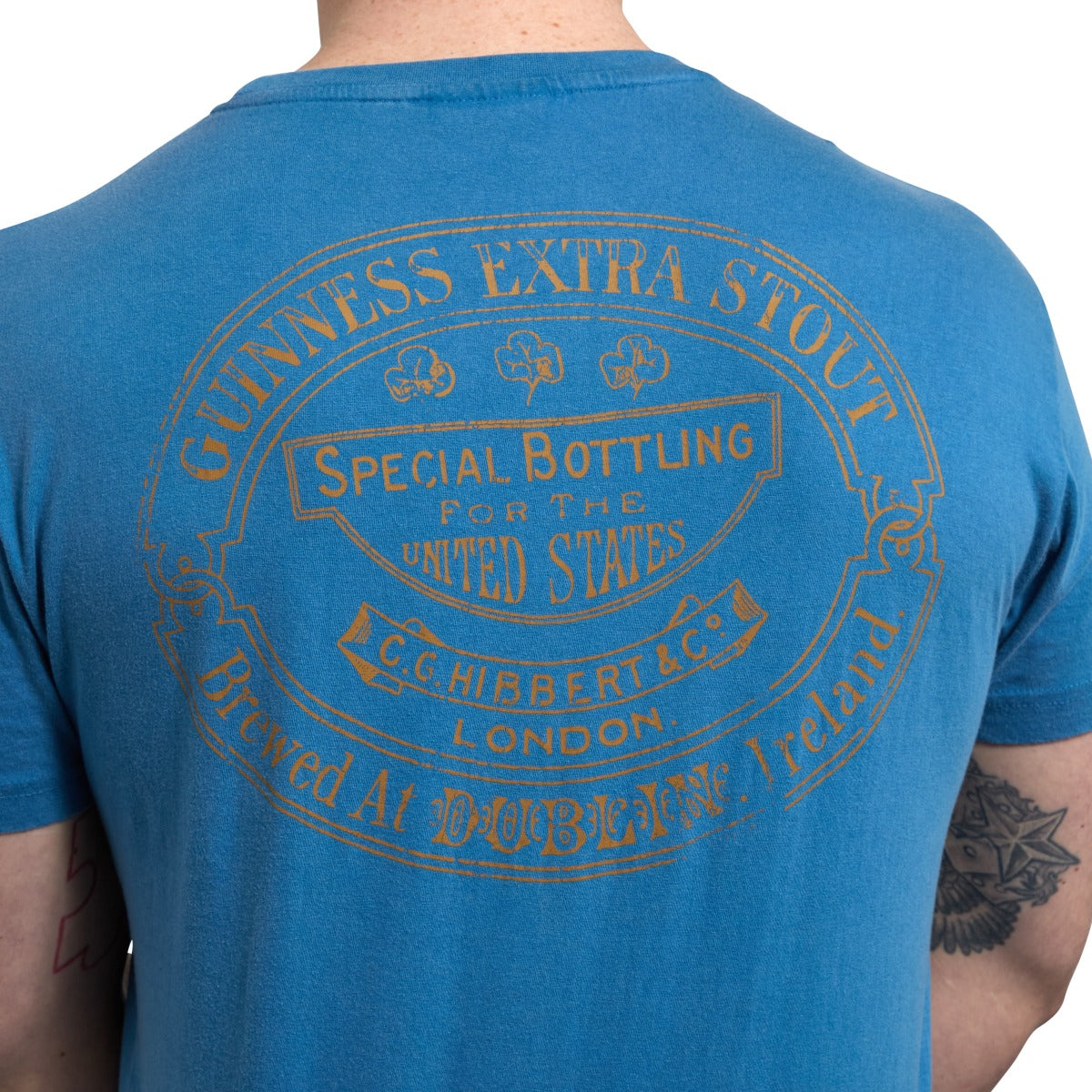 Back of a person wearing a Blue Guinness Harp Premium Tee with a large Guinness Extra Stout logo printed in faded orange text, indicating it as a special bottling for the United States by C.G. Hibbert & Co. Ltd., London. The vintage t-shirt proudly displays the iconic Guinness® Trademark Label.