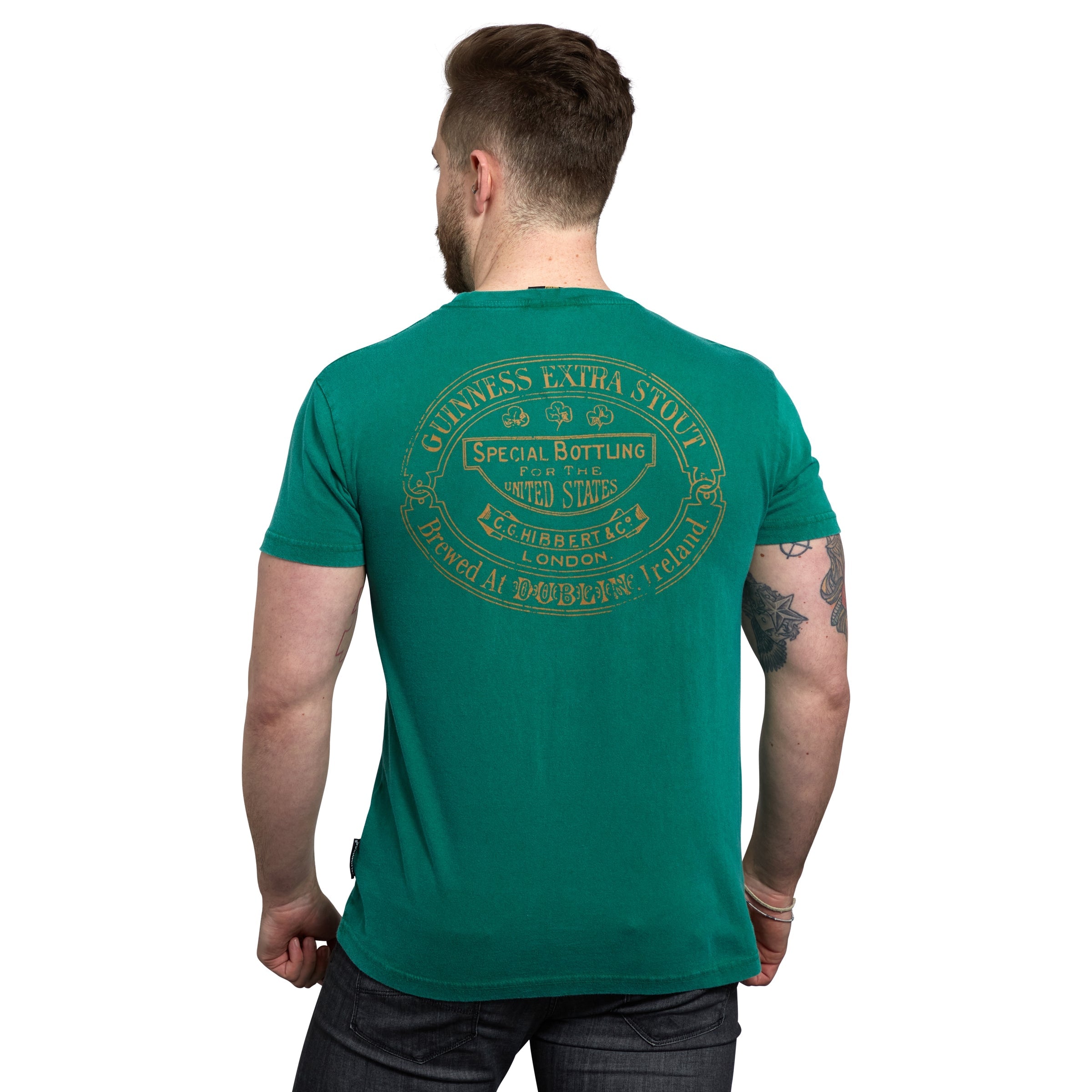 A man with tattoos is wearing a Green Guinness Harp Premium Tee from Guinness Webstore US, featuring vintage-style yellow text and designs on the back that read "Guinness Extra Stout." Perfect for any Guinness lover, the shirt has a loose fit for added comfort and style.